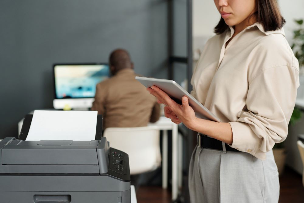 A young, professional woman stands in the foreground in front of a office copier while a colleague works in the background at a computer signifying Managed Print.