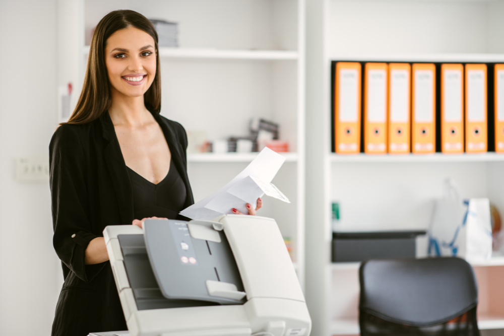 Smiling woman using a copier in an office, highlighting the benefits of managed print services. Shelves with organized files and office supplies are visible in the background.